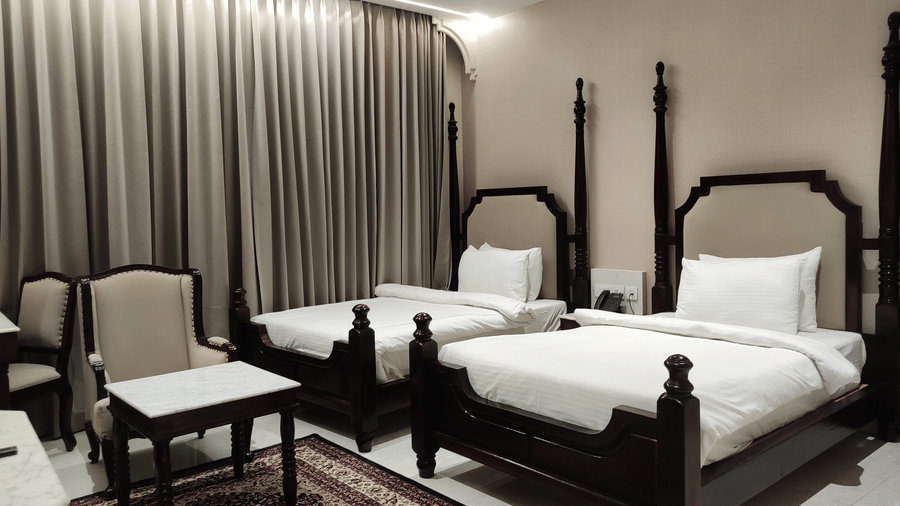 A bedroom at Anant Mahal, Jaipur featuring 2 double beds, a patterned rug, and long curtains covering a large window behind the seating area.