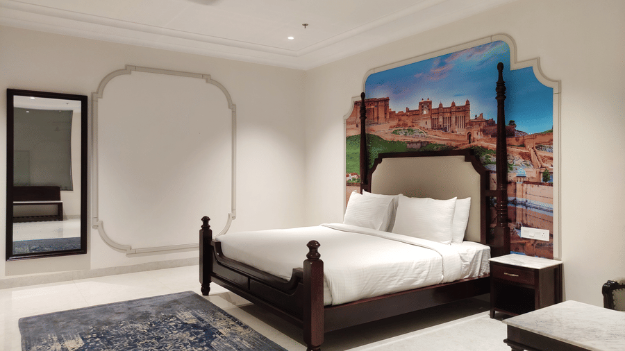 A wide shot of a bedroom at Anant Mahal, Jaipur featuring a king size bed, a blue patterned rug, a mirror, a ceiling fan, and a large mural behind the headboard.