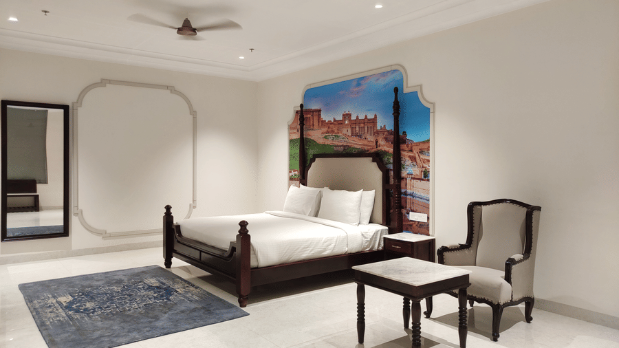 A bedroom view at Anant Mahal, Jaipur featuring a king size bed, a blue rug, a mirror, a seating area with a small table and a cushioned chair.