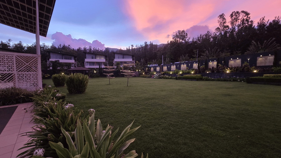 Green lawn at Bindiga Peak Resort surrounded by plants and structures under a pink evening sky.