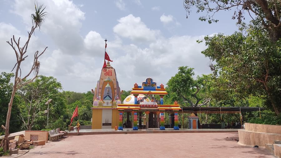 A serene temple painted in bright hues with a tall shikhara and flag, set in an open courtyard surrounded by trees under a partly cloudy sky.