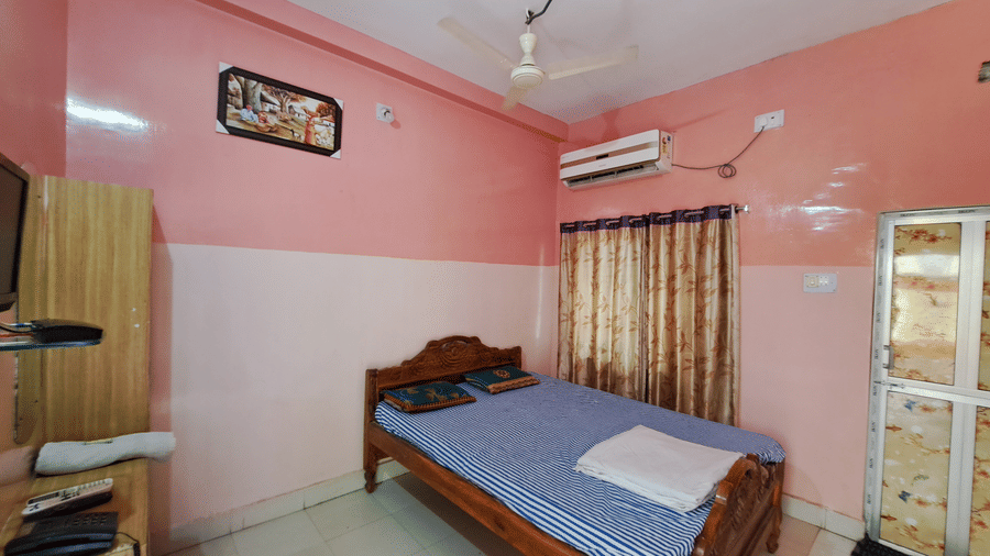 A Semi Deluxe AC Room at Khairi Resorts contains a bed, a ceiling fan, a wall-mounted AC unit, and a corner cupboard.