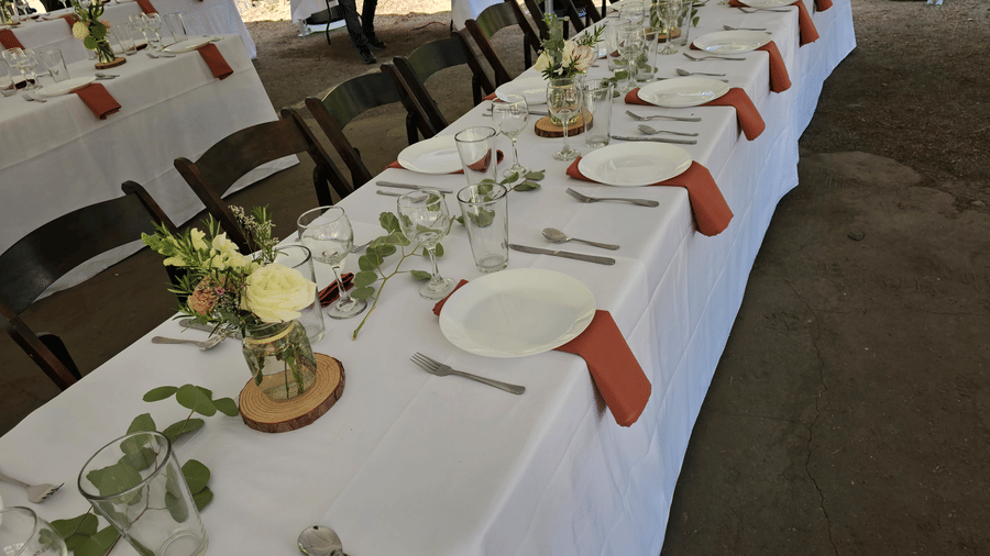 A long dining table at Kit Carson Lodge set with white linens, floral vases, and rust napkins under an outdoor pavilion