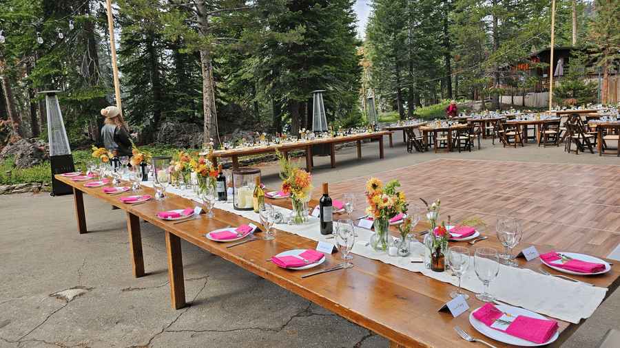 An outdoor dining setup at Kit Carson Lodge with a long wooden table decorated with bright floral center-pieces, napkins, and glassware, arranged on a stone patio surrounded by pine trees