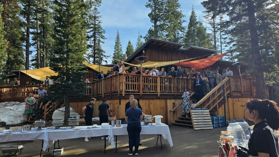 An outdoor gathering setup at Kit Carson Lodge featuring buffet tables and beverage stations on a paved area, with guests mingling near a wooden deck