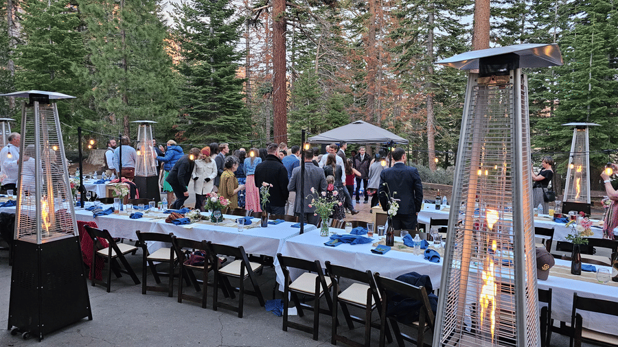 An outdoor dining setup at Kit Carson Lodge featuring long tables, chairs, and standing heaters surrounded by tall trees