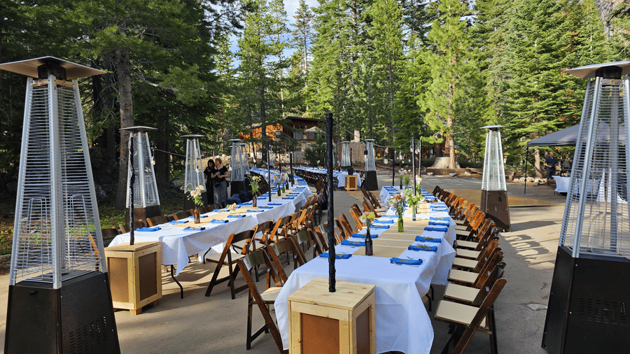 An outdoor dining area at Kit Carson Lodge featuring long tables covered with linens, surrounded by wooden chairs and tall outdoor heaters among pine trees