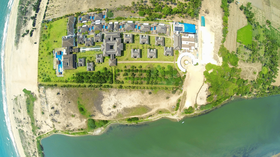 A high-angle aerial view showing the entire layout of the resort complex situated between a sandy beach and a wide river | Le Pondy
