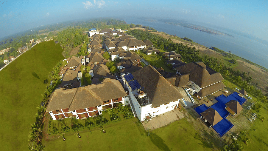 An aerial view of the resort complex featuring rows of villas with thatched roofs, a swimming pool, and lawns next to a body of water | Le Pondy