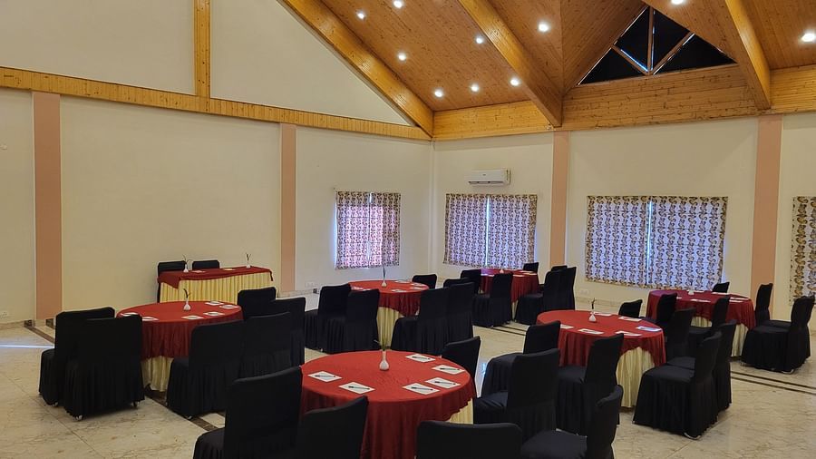 A spacious banquet hall with wooden ceiling beams, round tables, and elegant black and red seating at Dagshai Manor Kasauli - Inde Hotels.