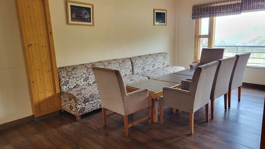A cosy seating area with a patterned sofa, wooden chairs, and large windows offering a scenic view at Dagshai Manor Kasauli - Inde Hotels.