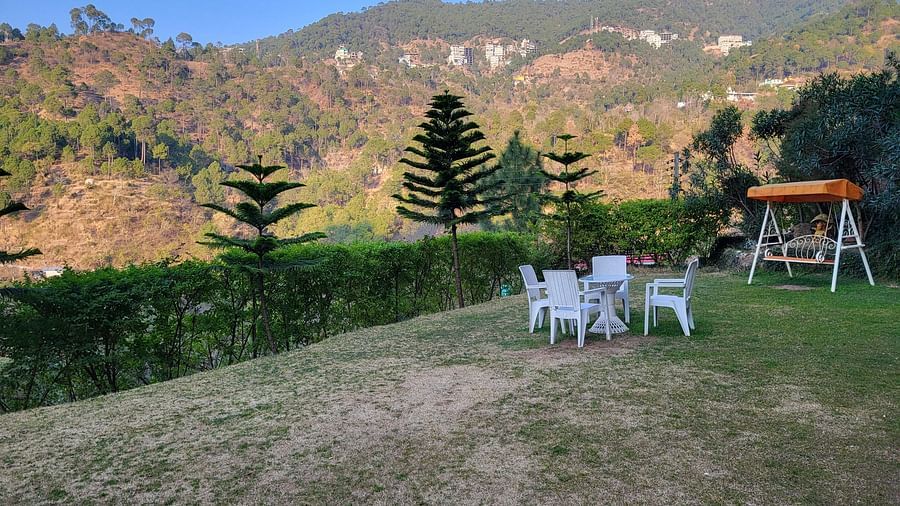 A scenic outdoor seating area with white chairs, a swing, and lush green surroundings overlooking the mountains at Dagshai Manor Kasauli - Inde Hotels.