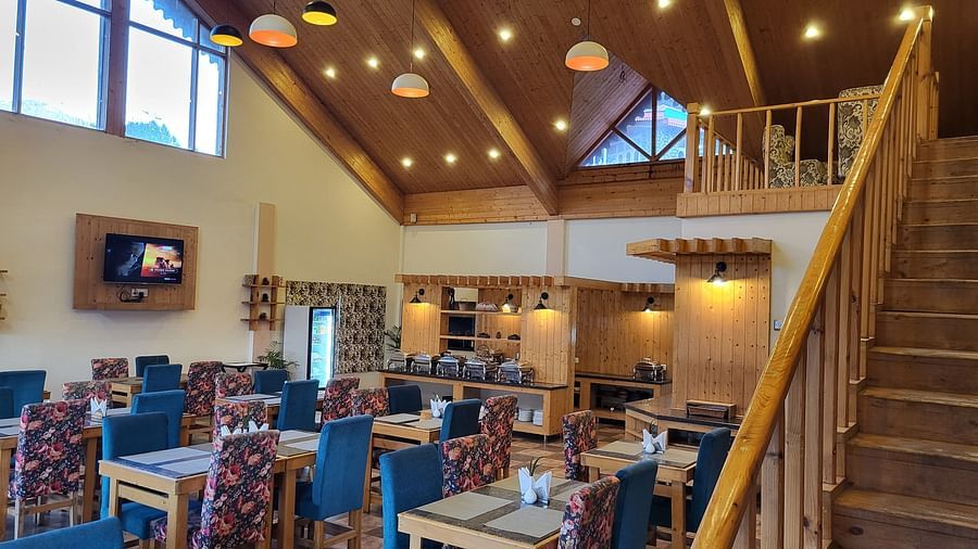 A well-lit, high-ceiling restaurant with wooden interiors, blue cushioned chairs, and an open staircase at Dagshai Manor Kasauli - Inde Hotels.