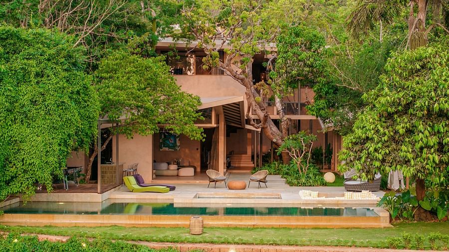 The front exterior of Villa Moira by V Escapes, featuring a sleek, long swimming pool and lush green lawns, surrounded by dense trees.
