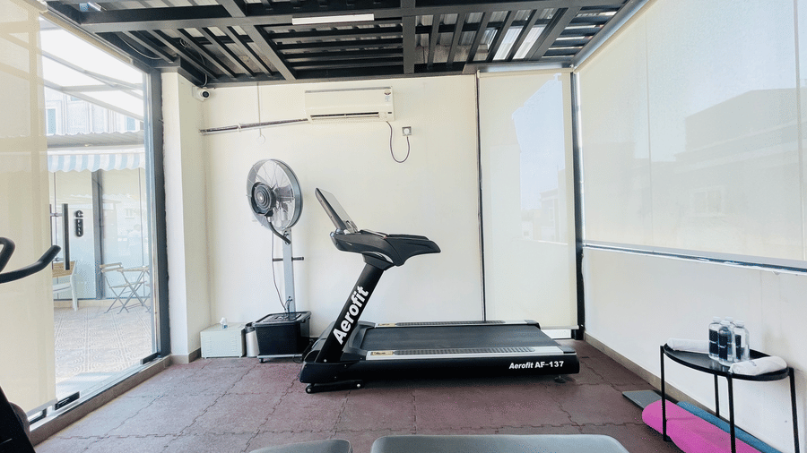 Open-air fitness area at Hotel Kyzen Smart Stays Madhapur featuring a treadmill, exercise bench, and standing fan with large glass walls and natural light.