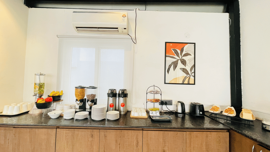  Corner beverage and condiment station at Hotel Kyzen Smart Stays Madhapur with juice dispensers, crockery, and framed artwork on the wall under dark ceiling beams.
