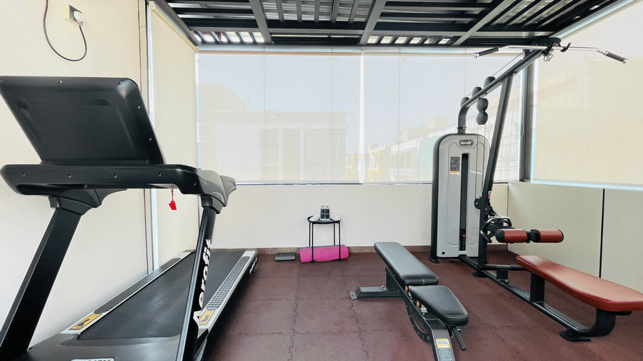 Rooftop gym at Hotel Kyzen Smart Stays Madhapur featuring a treadmill, exercise bench, and weight equipment surrounded by glass panel walls with natural daylight.
