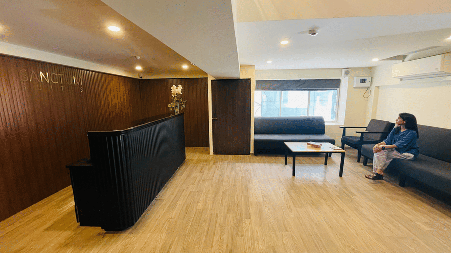 The reception and lobby at Sanctum Suites, Richmond Road, Bangalore features a black desk, a wooden coffee table, a window, and a person sitting on the sofa.