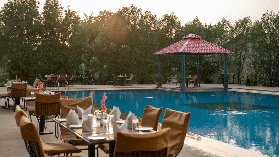 A close view of dining tables placed close to the clean swimming pool canopied by tall trees at Bamboo Saa Mulberry Resort, Pushkar.