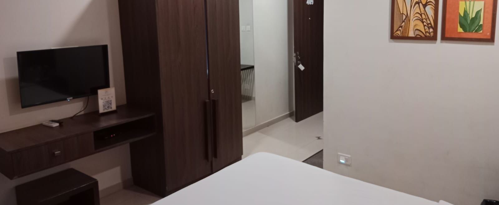 Full view shot of the Deluxe Room at Pravasa Ahmedabad featuring a wall mounted television beside a wooden wardrobe placed opposite to a double bed.