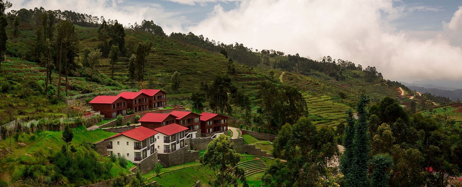 A drone shot of the GReaT Trails Kodaikanal By GRT Hotels property surrounded by lush greenery and trees with clouds hovering over