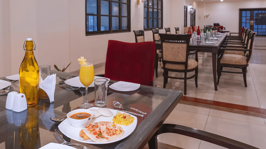 An indoor dining table set for a meal at Manorath Restaurant at Shri Vallabh Vilas Lords Plaza Nathdwara, with plated food, drinks , along with narrow dining table. 