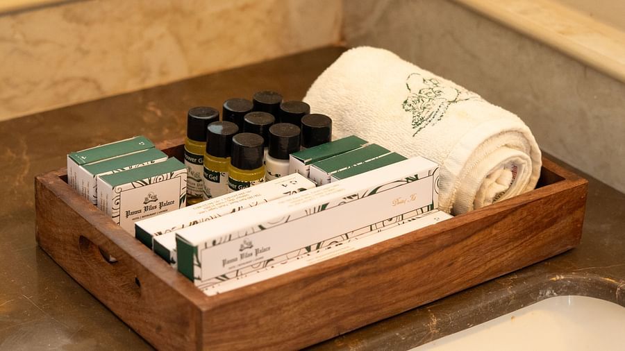 Wooden tray on a bathroom counter containing neatly arranged hotel toiletries including shower gel bottles, dental kits, soap boxes, and a rolled embroidered towel - Panna Vilas Palace