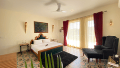 A bedroom with patterned floor tiles, a bed with a headboard, and an armchair. A ceiling fan, two wall sconces, a painting, two bedside tables, and large glass windows with curtains - Karma Lakelands