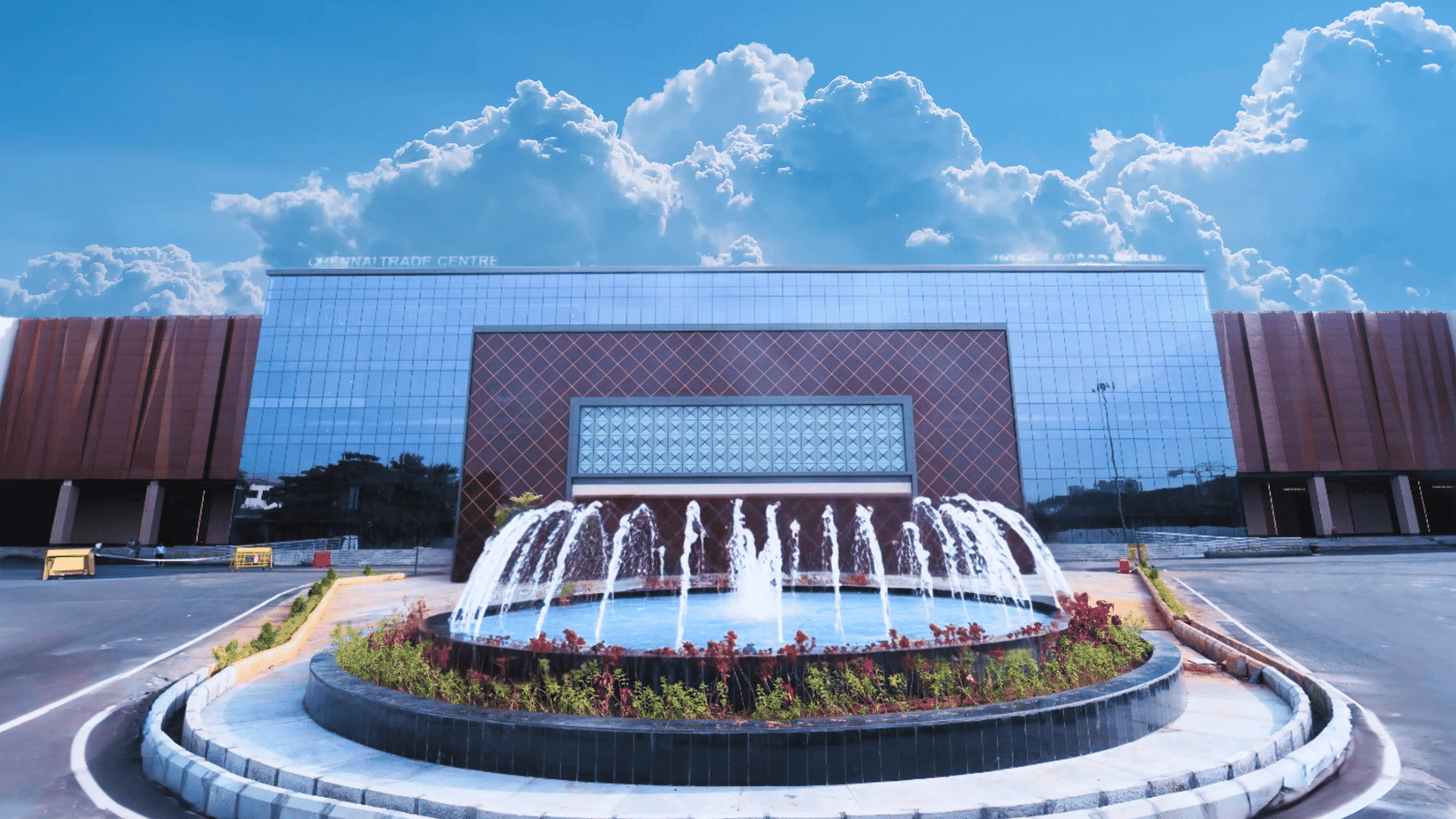 The modern, reflective glass facade of the Chennai Trade Centre, with a central fountain and a dramatic blue sky with white clouds overhead.
