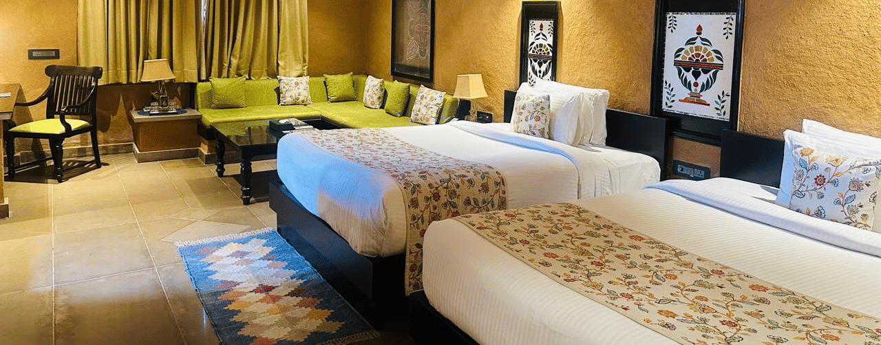 A spacious twin bedroom at Aamaghati Wildlife Resort featuring wooden ceilings, elegant lighting, comfortable twin beds, green seating and tasteful wall art.