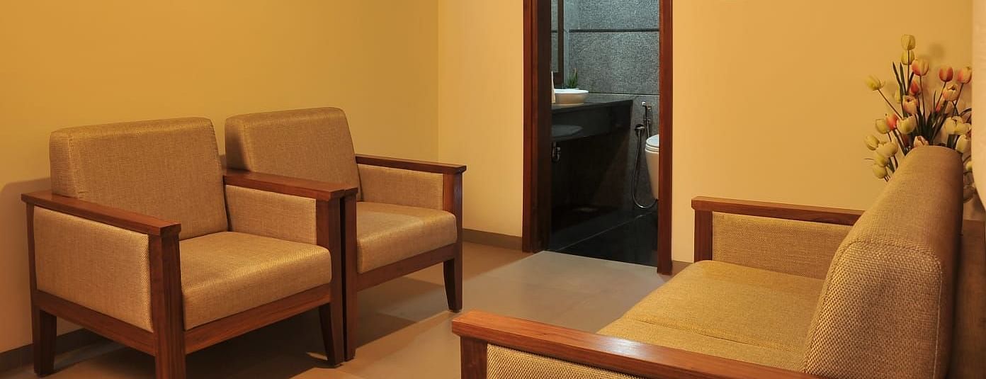 Side view of Suite's seating area at Dhanalakshmi Srinivasan Hotels, Perambalur shows two light brown couches with wooden frames facing each other, a wooden door, and a white vase with artificial tulip flowers.