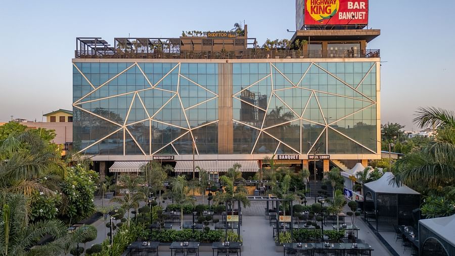 Exterior view of Hotel Highway King, Jaipur, with a large glass facade and rooftop signage, surrounded by greenery.