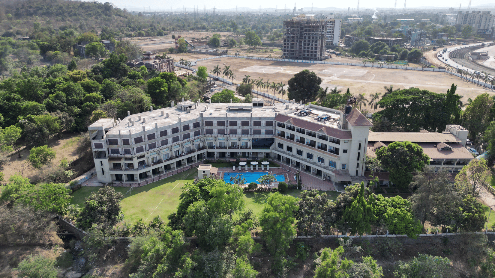 An aerial view of VITS Kamats Resort, Silvassa, a resort in Silvassa for weddings, surrounded by trees with a swimming pool, a lawn area in front of it and a highway, open land and an under-construction building in the background.