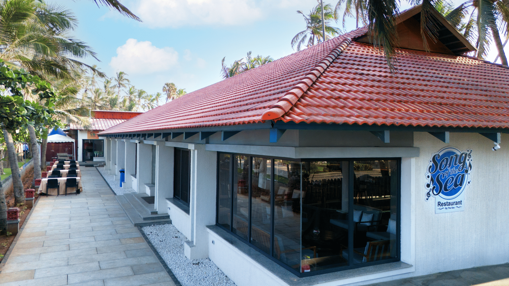 The exterior of the Song of the sea dining area, a single-story white building with a red tiled roof surrounded by tropical foliage at MGM Beach Resorts, ECR Chennai.