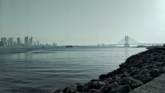 A rocky beach, with a sea bridge and buildings in the background.