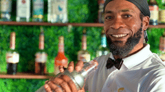 A smiling bartender holding a cocktail shaker in front of a colorful bar display at The Soco Hotel.