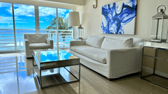 A stylish beachfront living room at The Soco Hotels, featuring modern furnishings, wall art, expansive windows, and a stunning ocean view.
