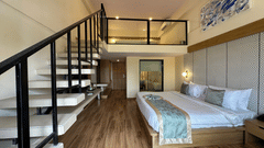 An overview of the Duplex Room with a king-size bed, a painting above the bed, side table, wall mounted lamps, a see through washroom, and a stairs leading to the second deck having an AC wall mounted at VITS Kamats Resort, Silvassa, the best resort in Sil