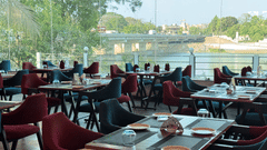 A close-up of the four chair seating with maroon and navy coloured chairs and a large glass panel overlooking the Ganga River and a bridge in view at the Riverside Restaurant in Silvassa inside VITS Kamats Resort, Silvassa.