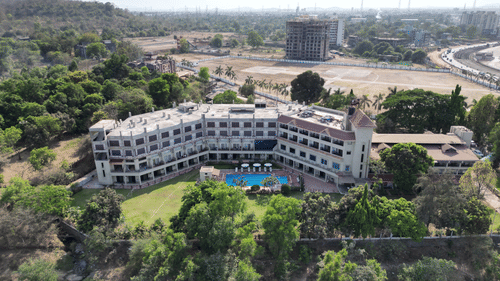 An aerial view of VITS Kamats Resort, Silvassa, a resort in Silvassa for weddings, surrounded by trees with a swimming pool, a lawn area in front of it and a highway, open land and an under-construction building in the background.