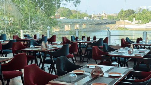 A close-up of the four chair seating with maroon and navy coloured chairs and a large glass panel overlooking the Ganga River and a bridge in view at the Riverside Restaurant in Silvassa inside VITS Kamats Resort, Silvassa.