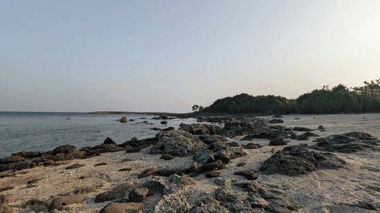 A rocky shore with a calm sea and a small island under a hazy sky.