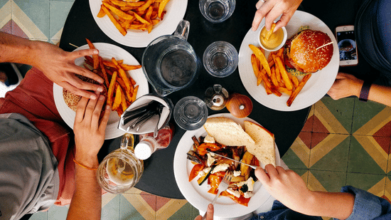 a group of four people eating pasta, burger and fries on a plate along with a beer glass on the table besides a water jug  and glasses 