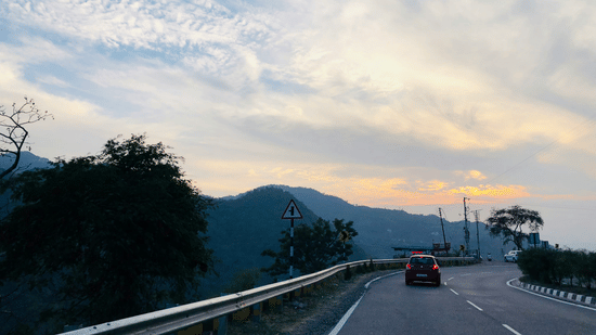 Scenic mountain road in Kasauli at sunset, with a car driving along a winding highway surrounded by rolling hills and a colourful evening sky.