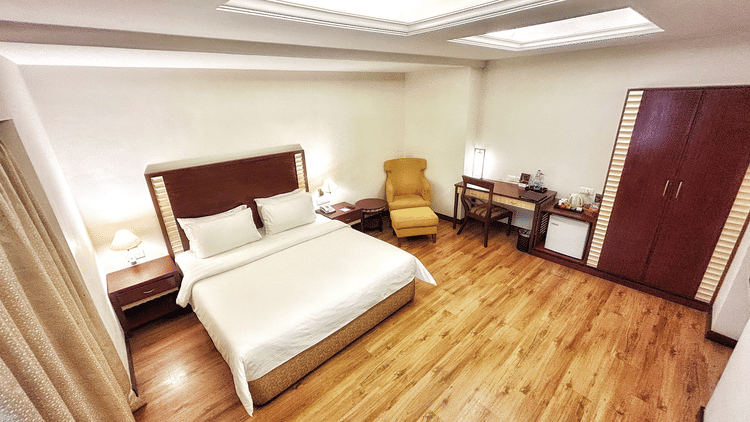 Deluxe Room at The Residency Karur with wooden flooring, a double bed, cosy armchair, work desk, warm lighting, and minimalist modern decor.