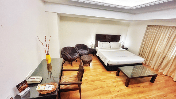 Residency Suite at The Residency Karur shows a double bed, a glass coffee table, two round chairs, and a desk with a wooden chair on a wooden floor.