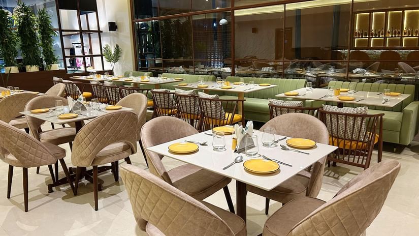  A modern indoor restaurant dining area at Amer Greens featuring square tables with soft chairs in the foreground and a long bench seat along the large window wall in the background.