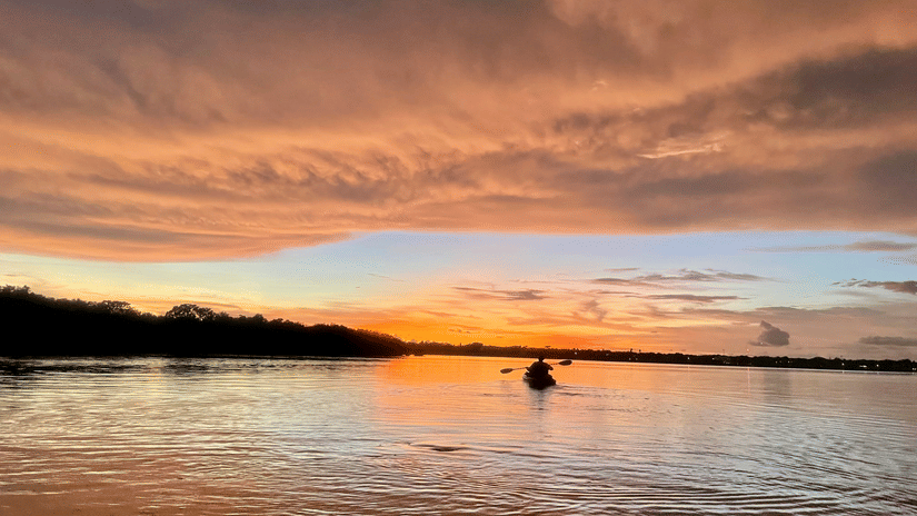 A view from the shore of a beach of a person night kayaking in Havelock on the waterbody with hills in the distance and different hues in the sky.