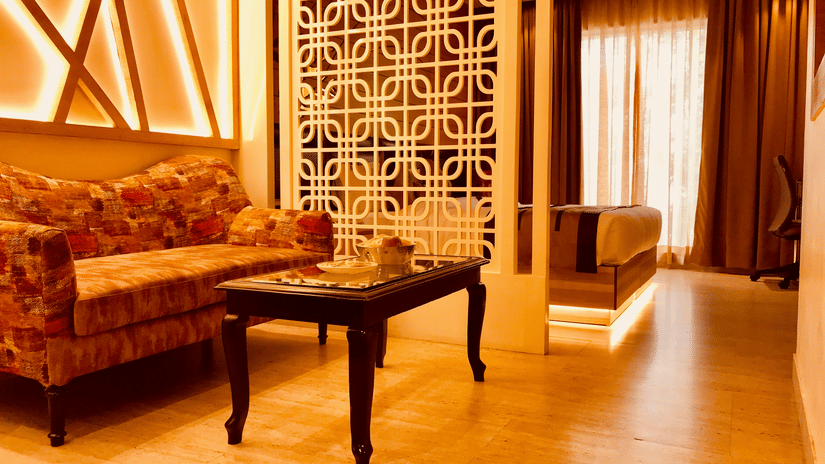 A seating area at a guest room at JRD Hotels showcasing multi-seating sofa, a coffee table, wooden flooring, and a double bed at the backdrop.