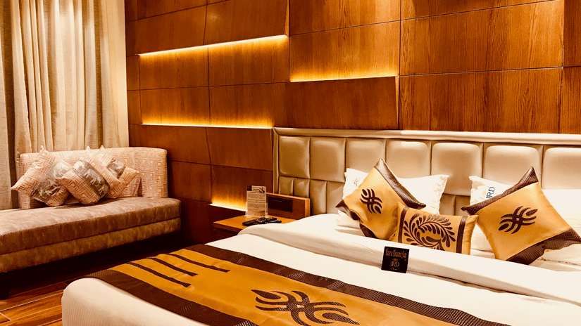 The Executive hotel at JRD Pride showcasing a double bed, a multi-seating sofa, wooden flooring, and ceiling lights.